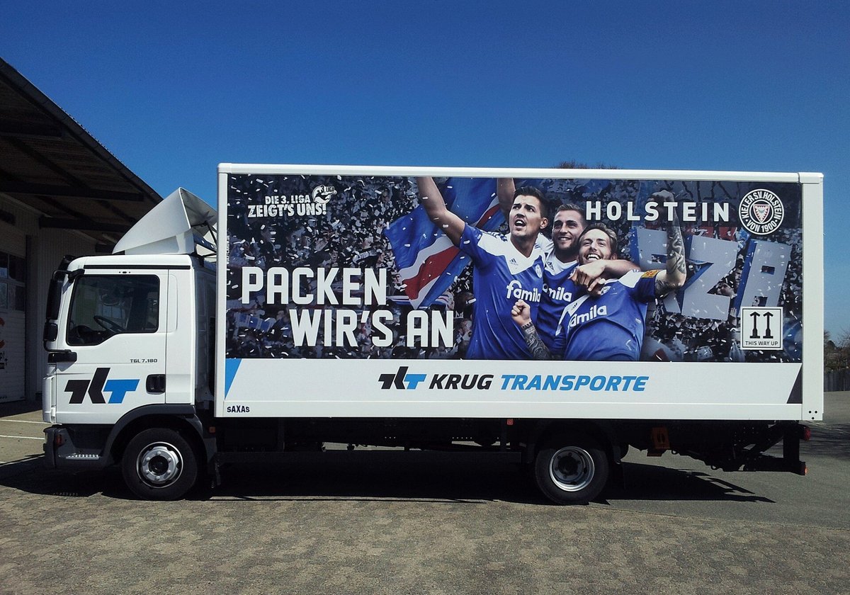 LKW mit auffälliger Werbebeschriftung von Krug Transporte, featuring Holstein Kiel Fußballspieler und dem Slogan „Packen wir’s an“, umgeben von jubelnden Fans.